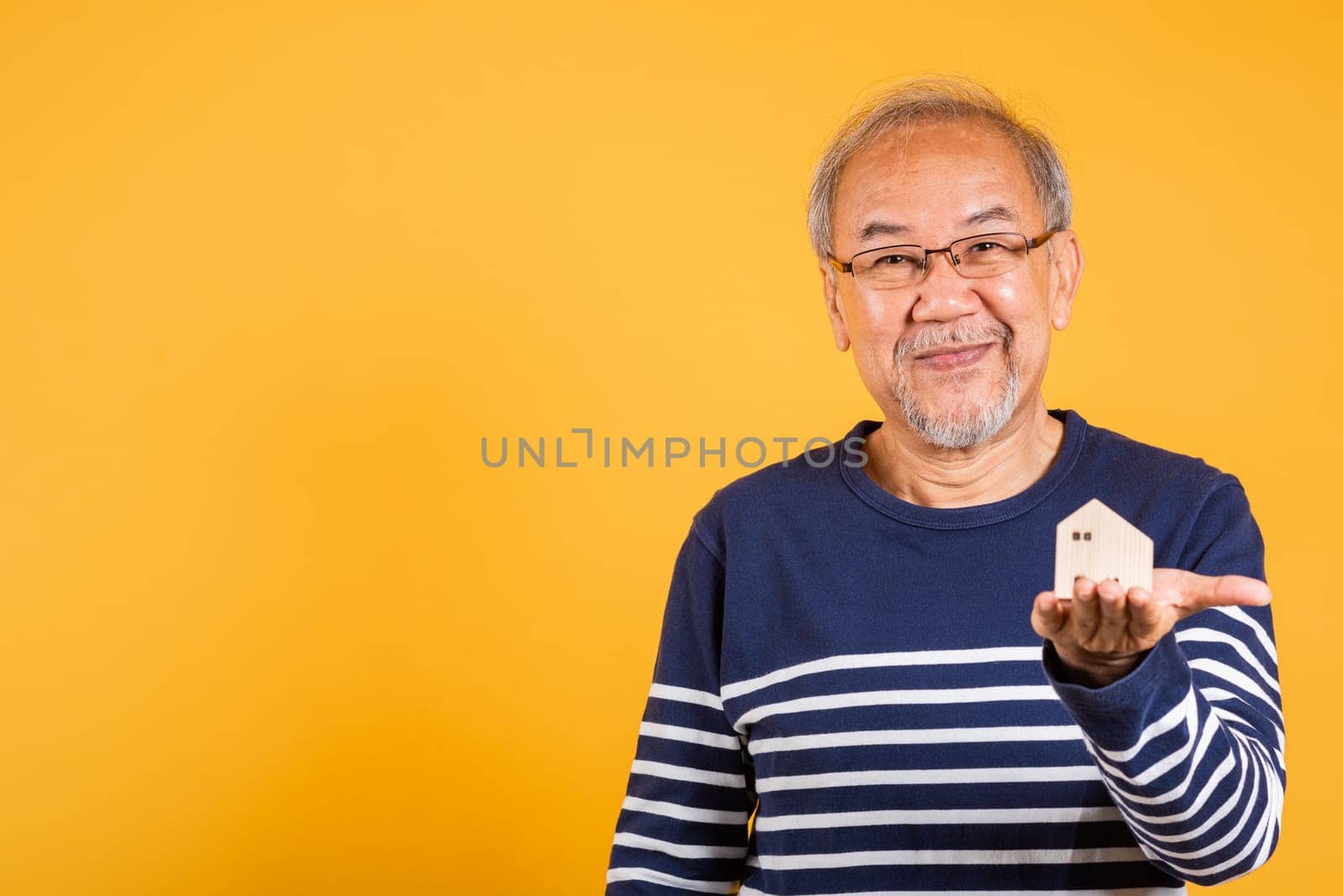 Portrait Asian smiling old man with glasses holding small house model studio shot isolated yellow background, happy senior man pensioner with grey hair have home property