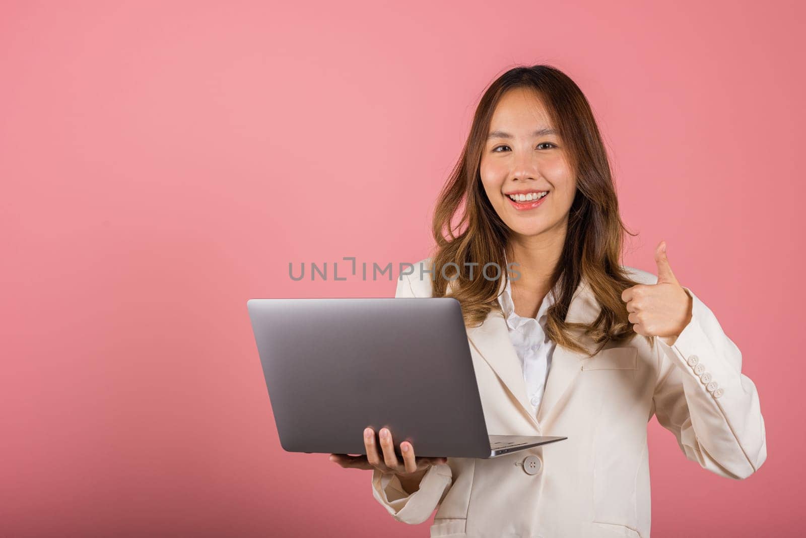 Portrait of happy Asian beautiful business woman confident smiling face holding using laptop computer and showing thumb up for like gesture, studio shot with copy space isolated on pink background