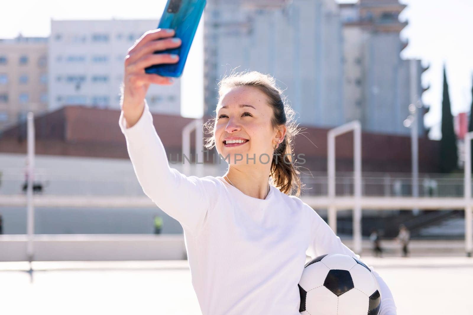 smiling young caucasian woman taking a selfie photo with mobile phone while holding a soccer ball, concept of social media and football lifestyle