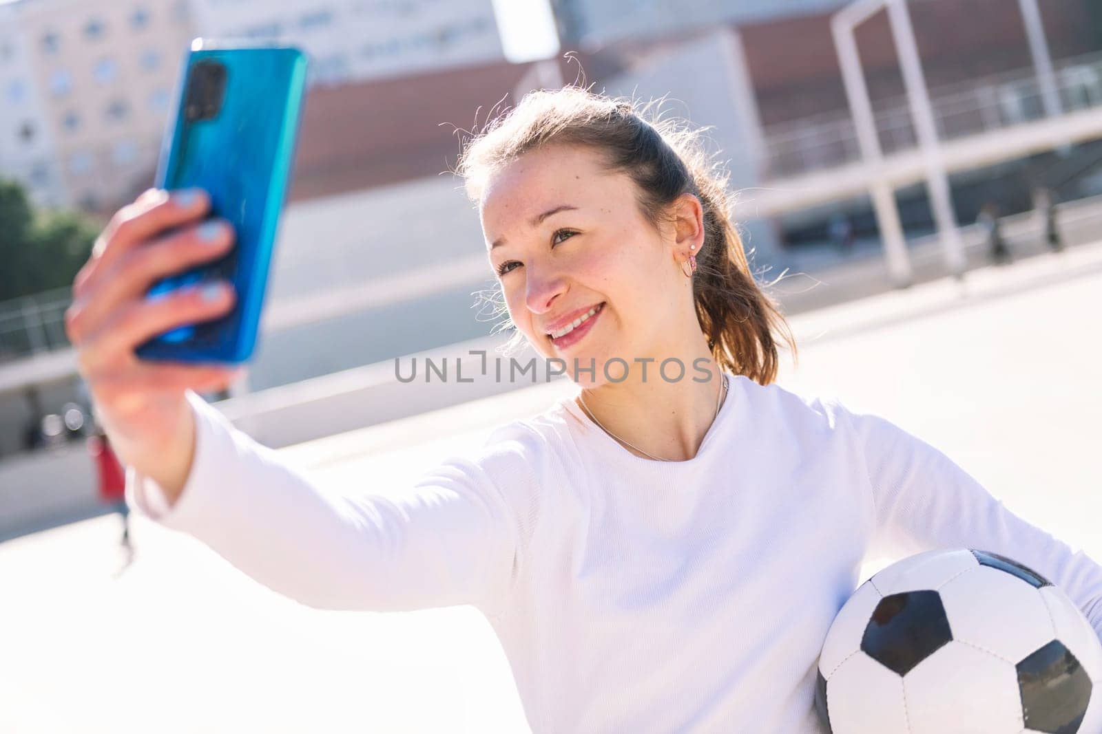 smiling young caucasian woman taking a selfie photo with mobile phone while holding a soccer ball, concept of social media and football lifestyle