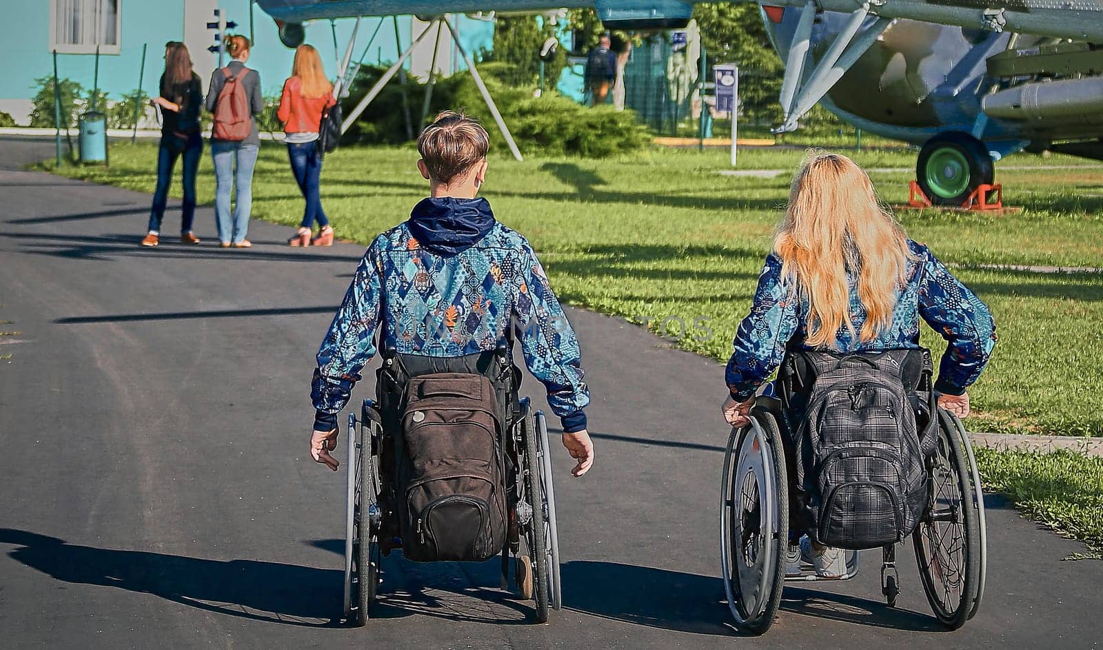 Two individuals, a man and a woman, both in wheelchairs, are exploring an outdoor museum featuring retired military helicopters on a sunny day, showcasing accessibility and inclusion.