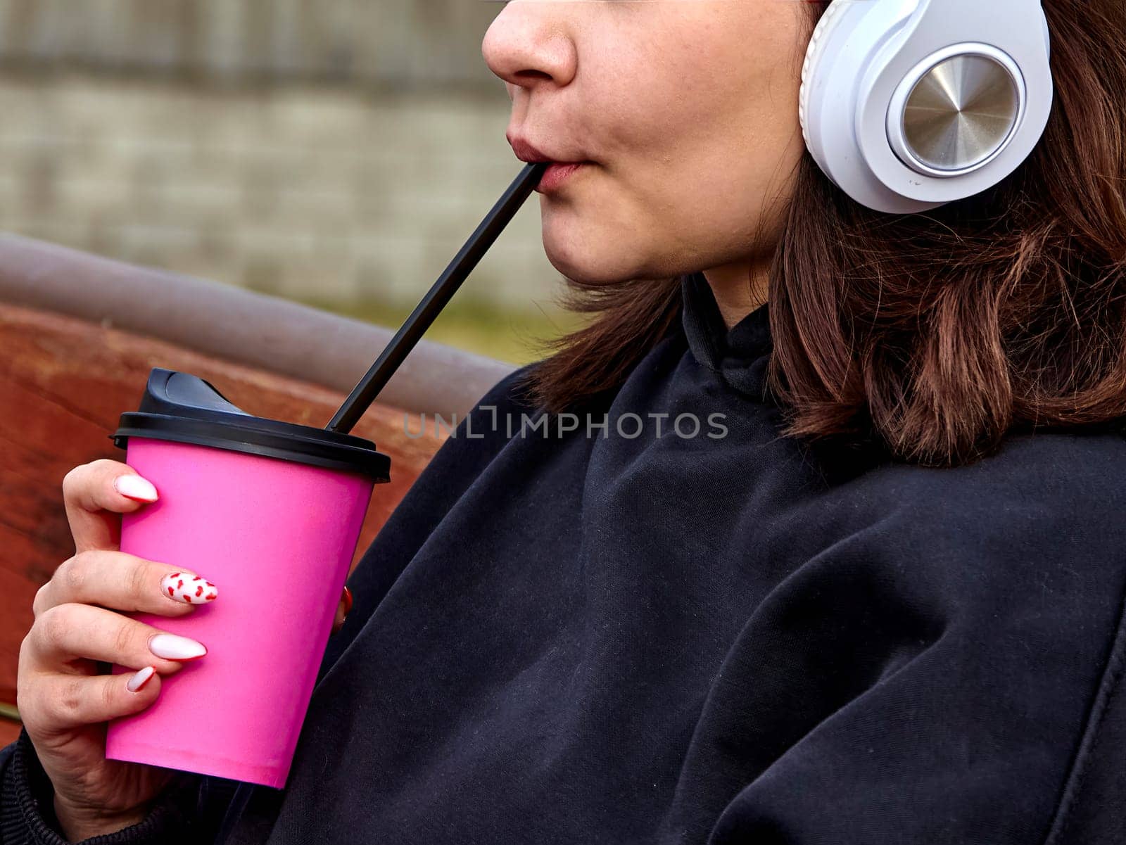 A young woman with long brown hair and white headphones is drinking from a pink cup with a straw. She is wearing a black sweatshirt and has her nails painted pink with white polka dots.