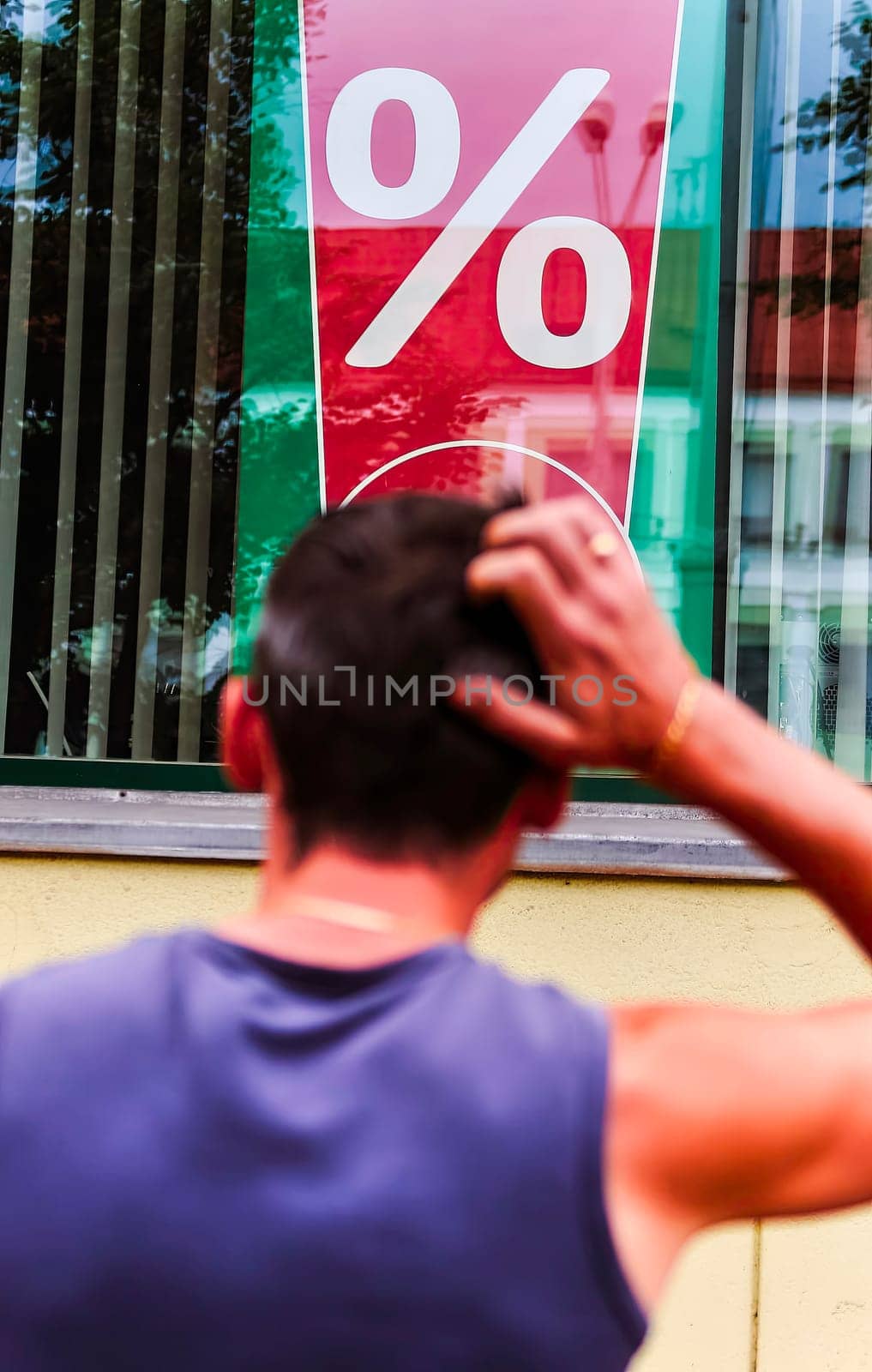 A man is standing outside a store, scratching his head as he observes a large percentage sign displayed in the window. His posture suggests he might be puzzled or considering the implications of the advertised discount, sale, or promotion.