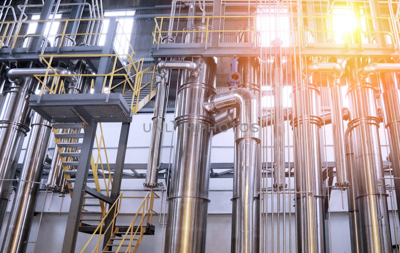 A chemical plant features a complex network of stainless steel pipes and glass vessels, creating an intricate system of interconnected straight and curved components in various sizes and shapes.