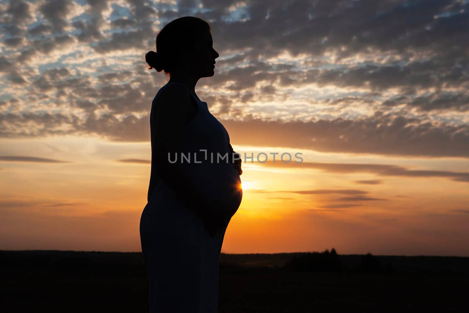 The silhouette of a pregnant woman at sunset with her hands on her belly. The sky is ablaze with color, and the clouds are forming a beautiful pattern. A truly stunning and peaceful scene.