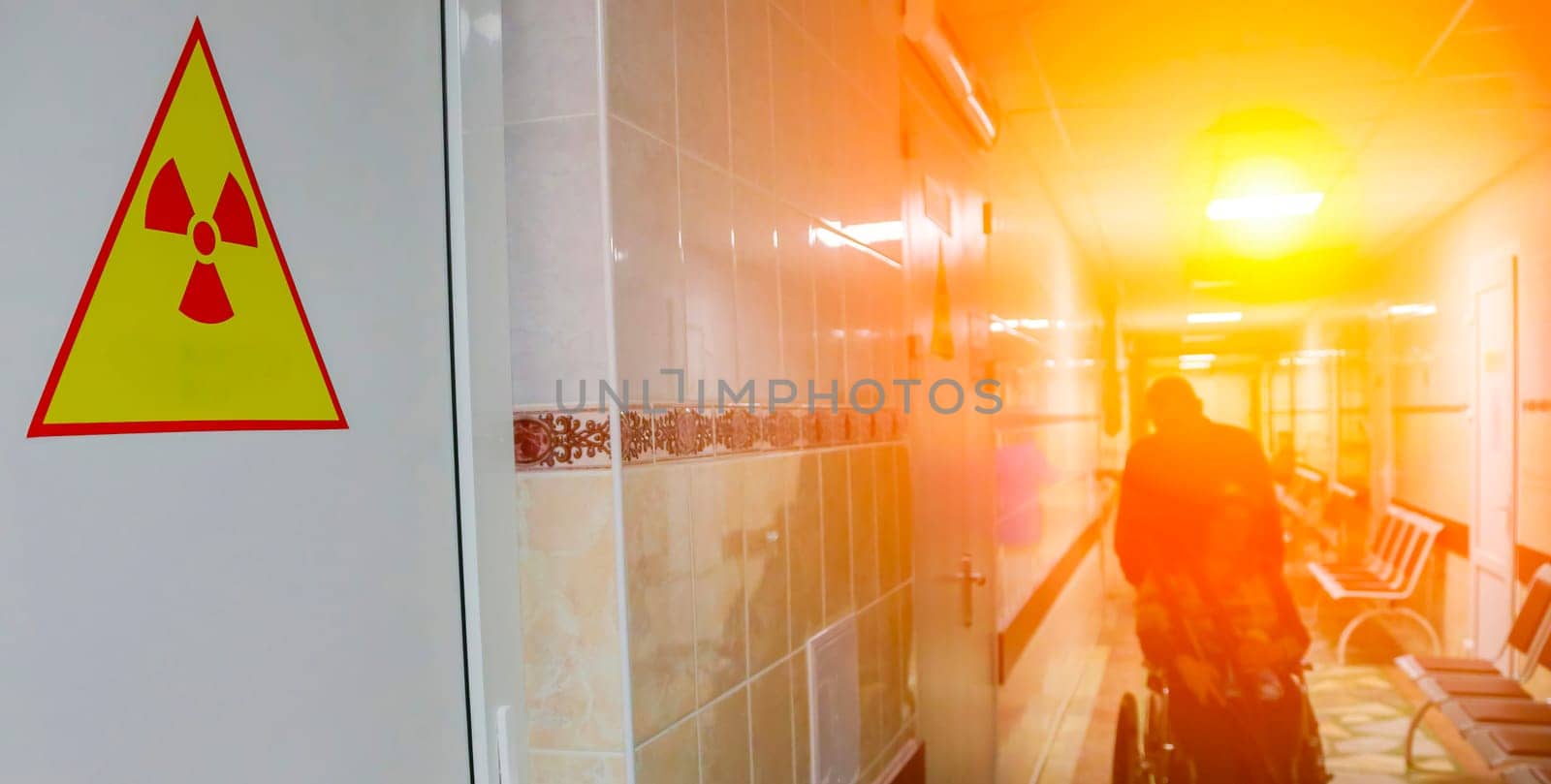 A person in a wheelchair pushes through a hospital corridor with a bright light illuminating the way. A yellow warning sign with a red radiation symbol is prominently displayed on the wall, highlighting the stark contrast between danger and hope.
