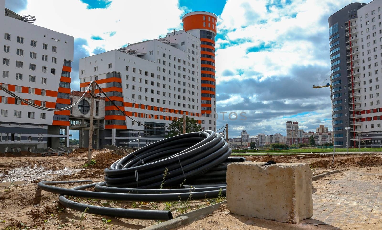 A pile of black pipes sits in front of two unfinished apartment buildings, with a concrete block in the foreground. Construction cranes are visible in the background, while a cityscape of other buildings stretches out under a partly cloudy sky.