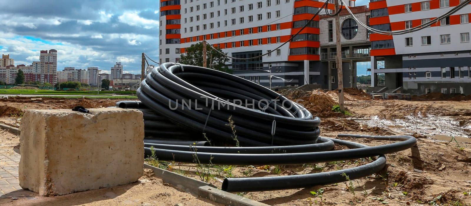 A large coil of black pipes sits on a dirt construction site, resting against a concrete block. The pipes appear to be part of a new infrastructure project, with a newly constructed apartment building in the background.