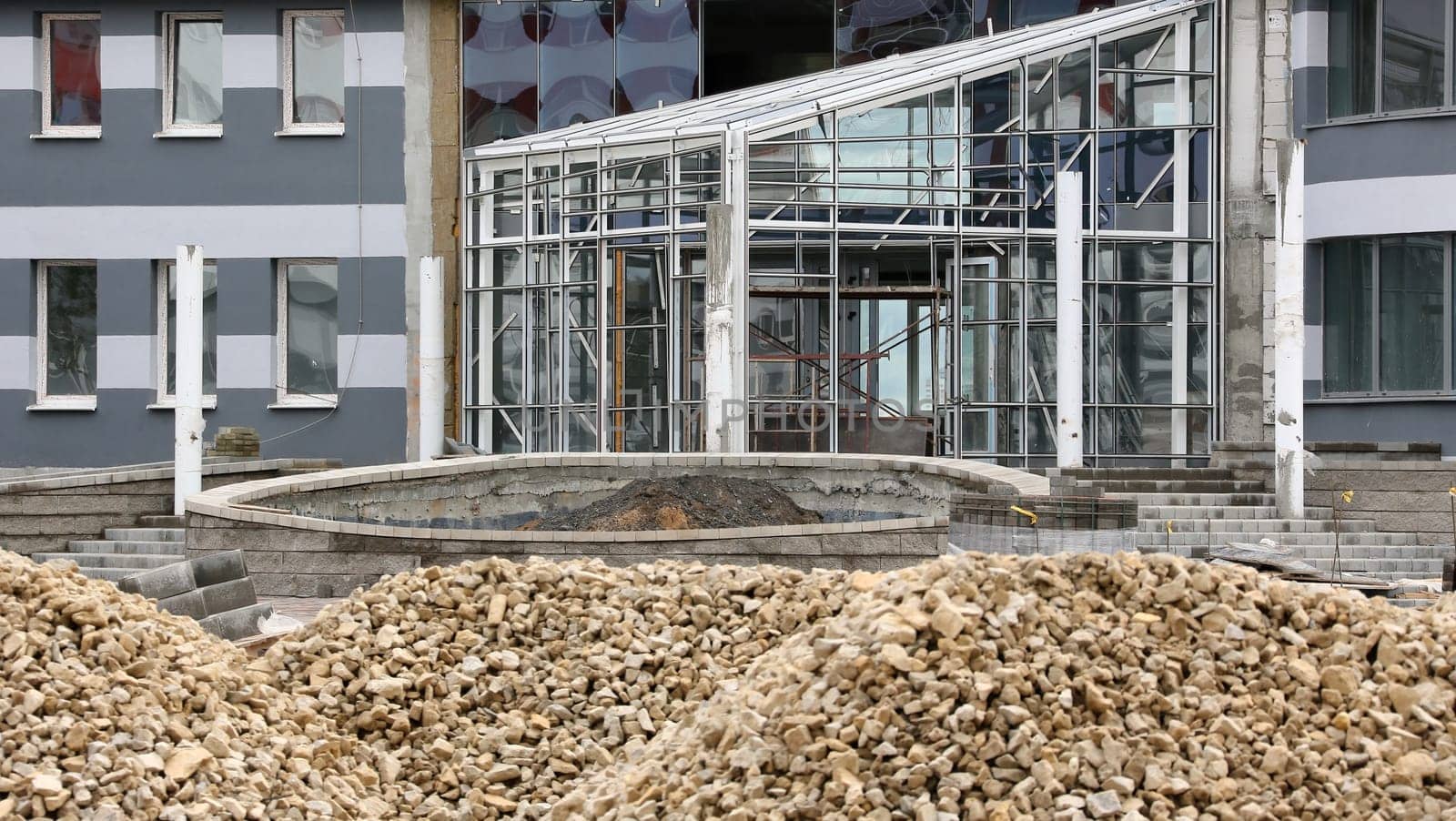 A modern building under construction with a large glass facade. The structure is incomplete with visible scaffolding and piles of construction aggregate in the foreground.