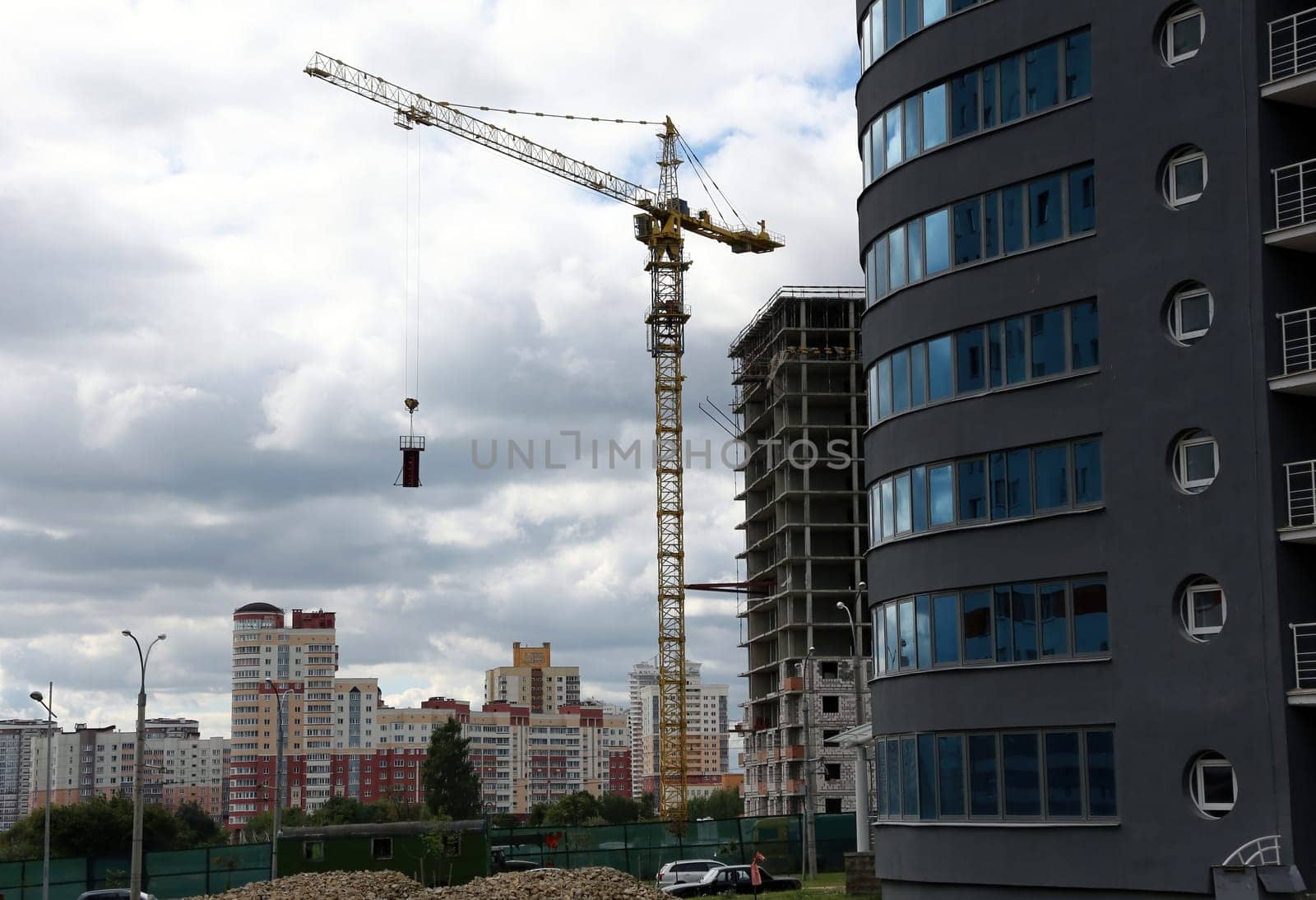 A construction crane stands tall against a cloudy sky, lifting a load of concrete as it builds new apartments in a bustling city.