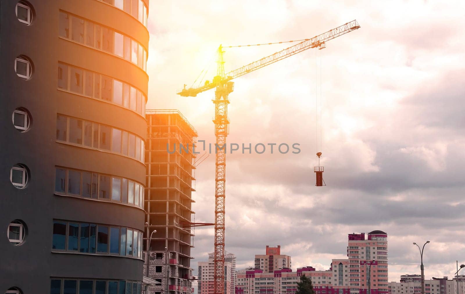 A construction crane stands tall against a backdrop of a city skyline, silhouetted by the setting sun. Buildings are under construction, showing the progress of urban development.