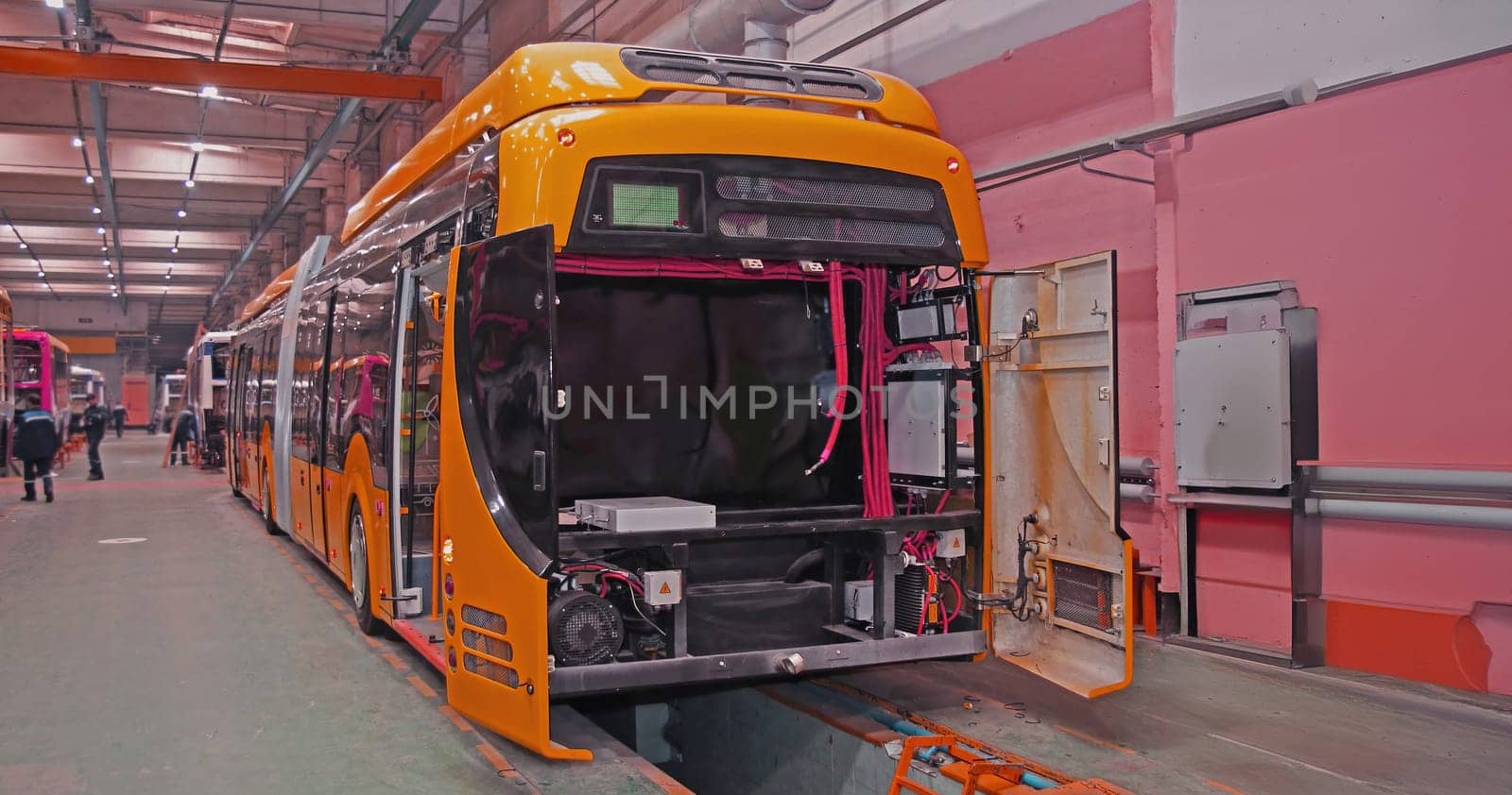 A bright orange electric bus is being assembled in a factory, with its doors open and various wires and components visible. The bus is positioned on a platform with other buses behind it, creating a sense of production and progress.