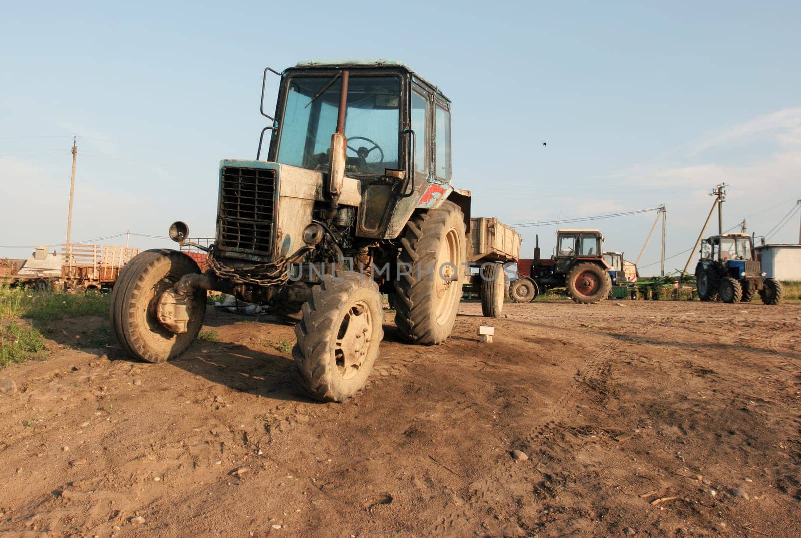 A weathered tractor sits in a rural field under a clear blue sky. The tractor is covered in rust, and its tires are worn down. The surrounding field is covered in dirt and grass, and there are other tractors in the distance.