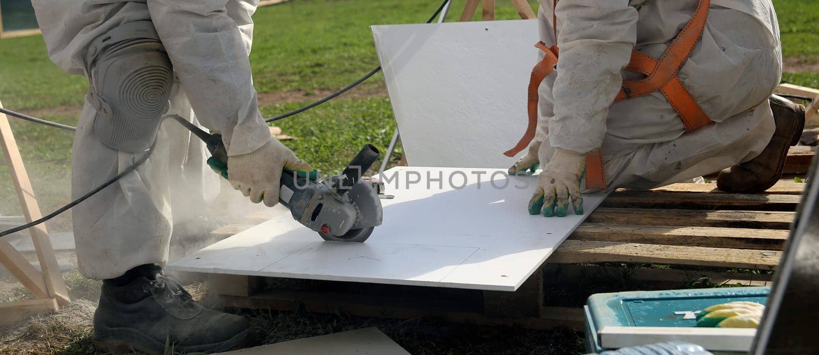 A construction worker in white overalls cuts a white panel with a grinder on a wooden pallet. The worker is wearing safety gloves and a harness.