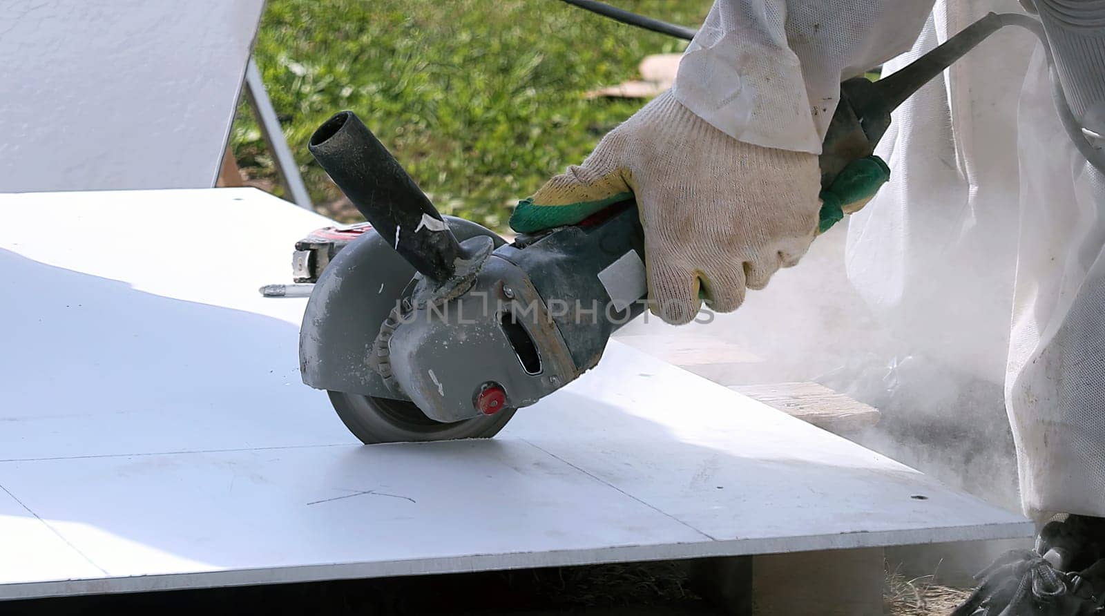 A person wearing gloves uses an angle grinder to cut through a white material outdoors. Dust and sparks fly up as the grinder spins.