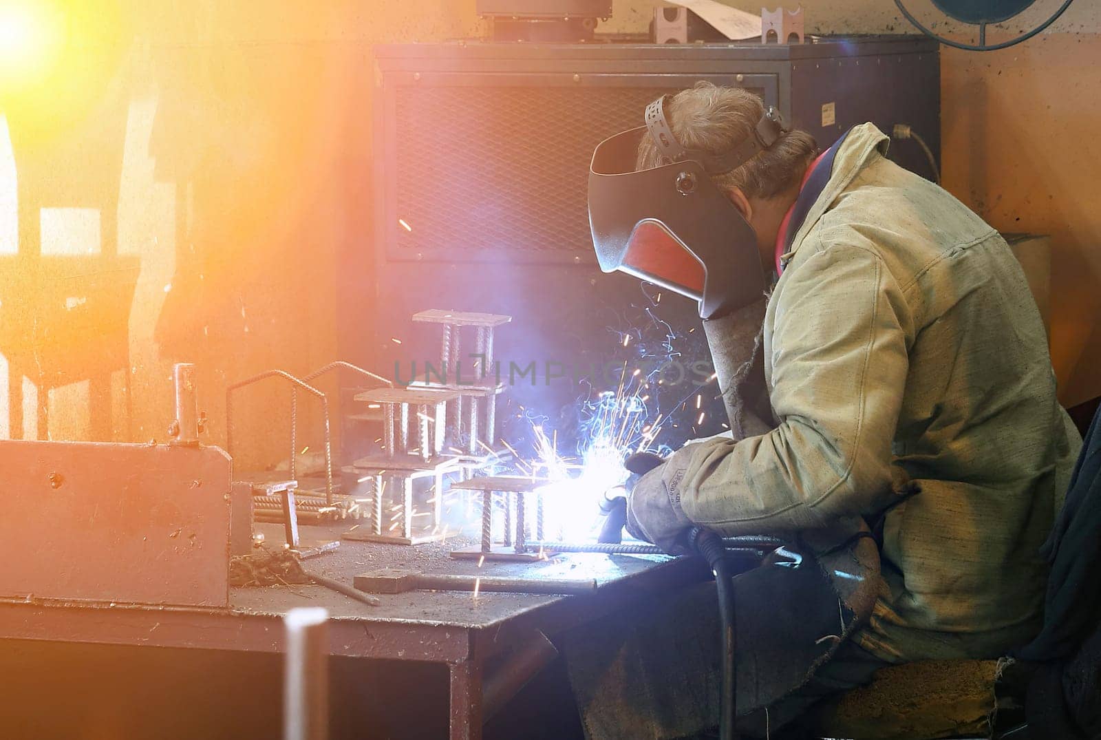 A welder works diligently, wearing a protective mask and gloves, amidst a shower of sparks as they skillfully weld metal pieces. The intense light of the welding torch illuminates the workshop, highlighting the workers concentration.