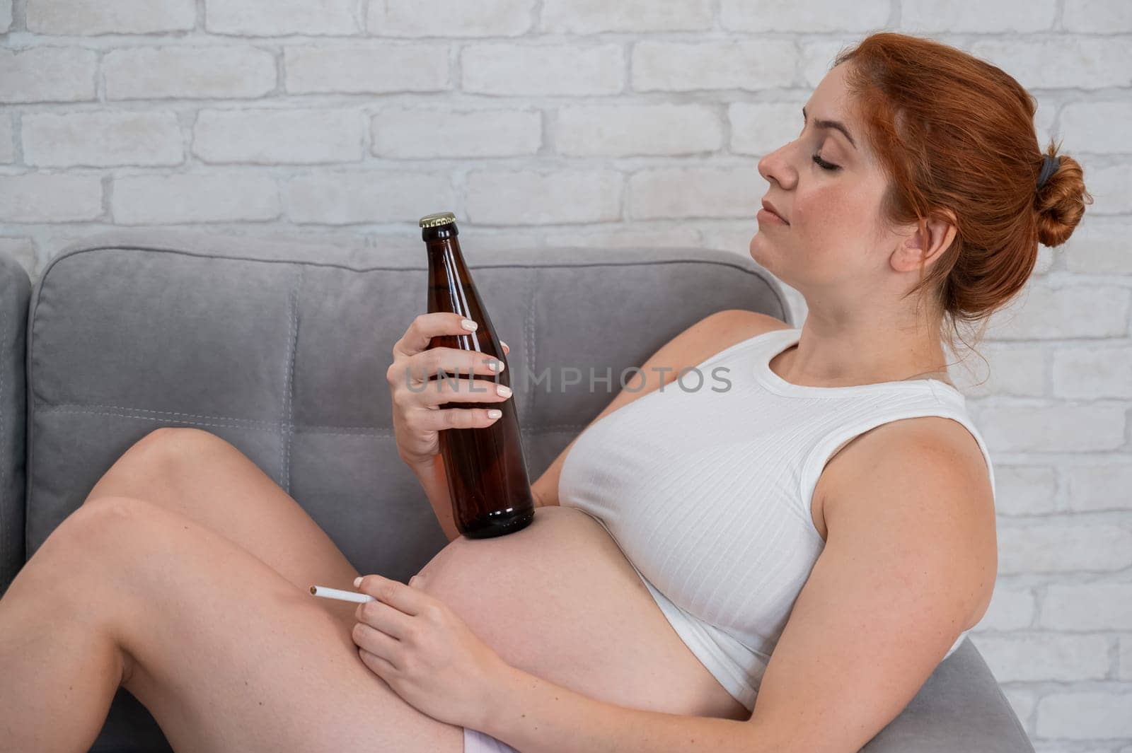 Red-haired pregnant woman drinks beer and smokes a cigarette while sitting on the sofa