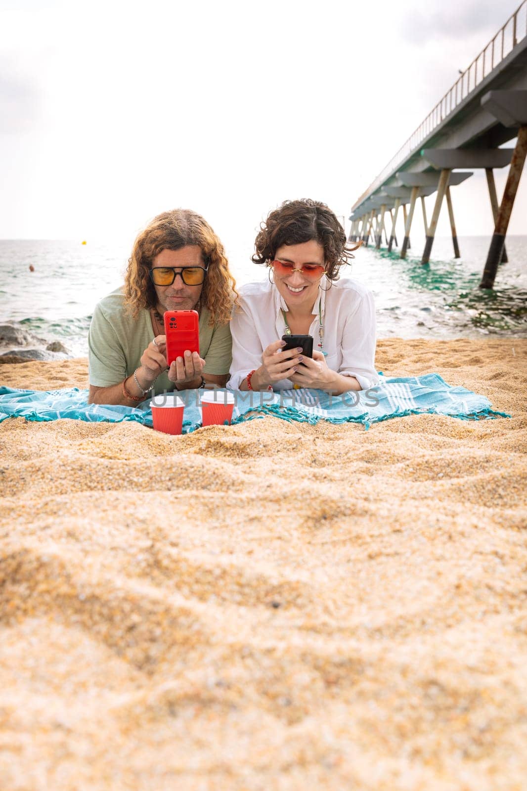 Middle-aged man and woman lying on the beach with surprised face, browsing smartphone apps. Concept: Holidays and Technology