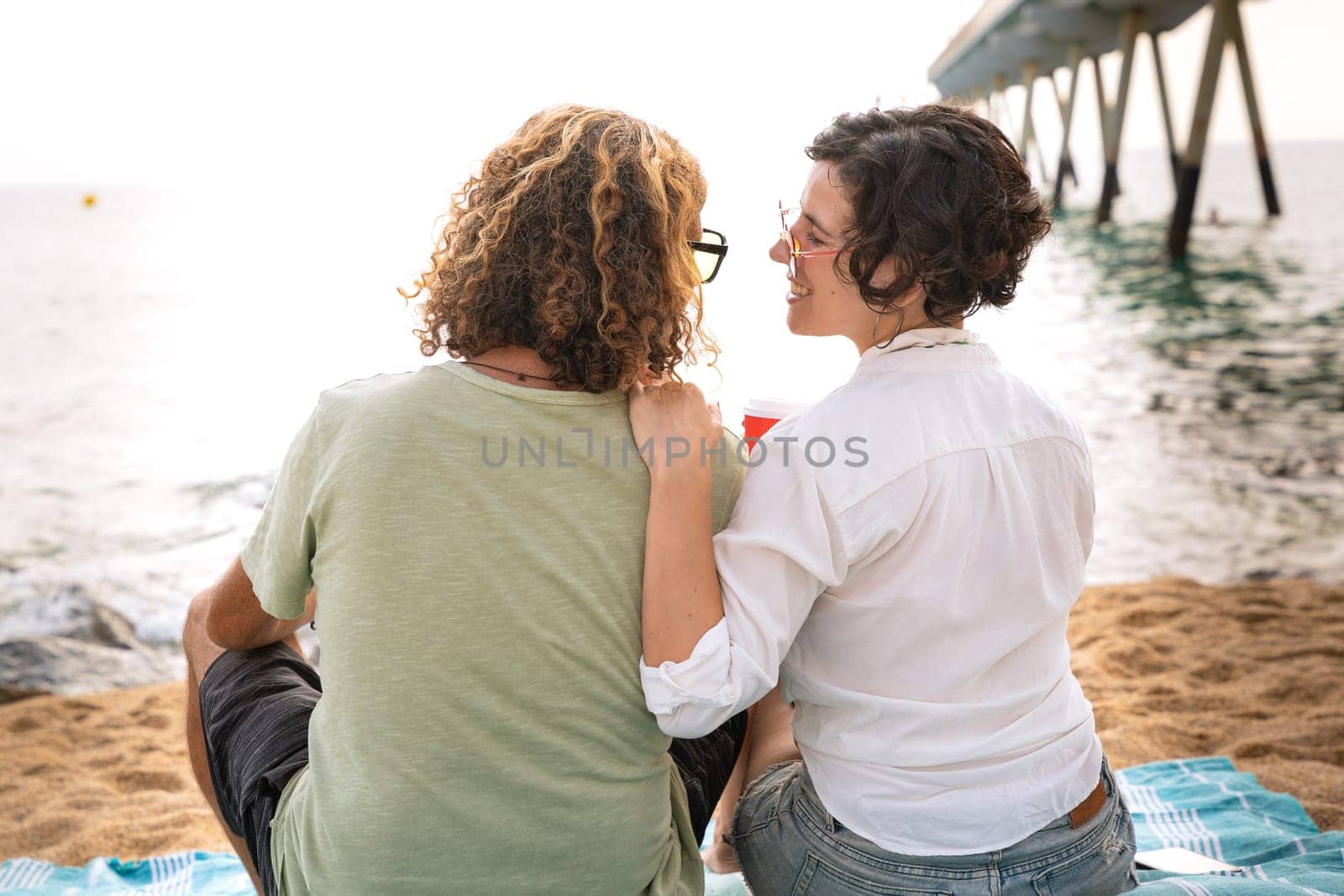 Rear view two happy friends sitting on the beach on vacation,looking at the sea. by mariaphoto3