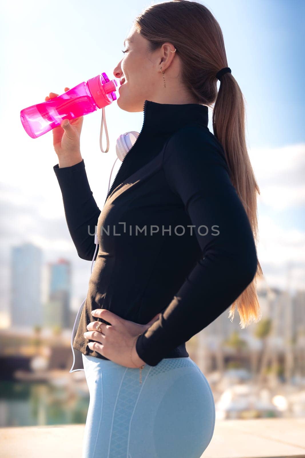 Side view runner girl drinking water from a sports bottle outdoors.Vertical by mariaphoto3