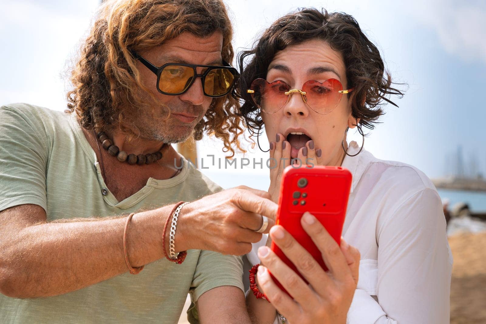 Middle-aged man and woman sitting on the beach with surprised face, browsing smartphone apps. Concept:Holidays y Technology