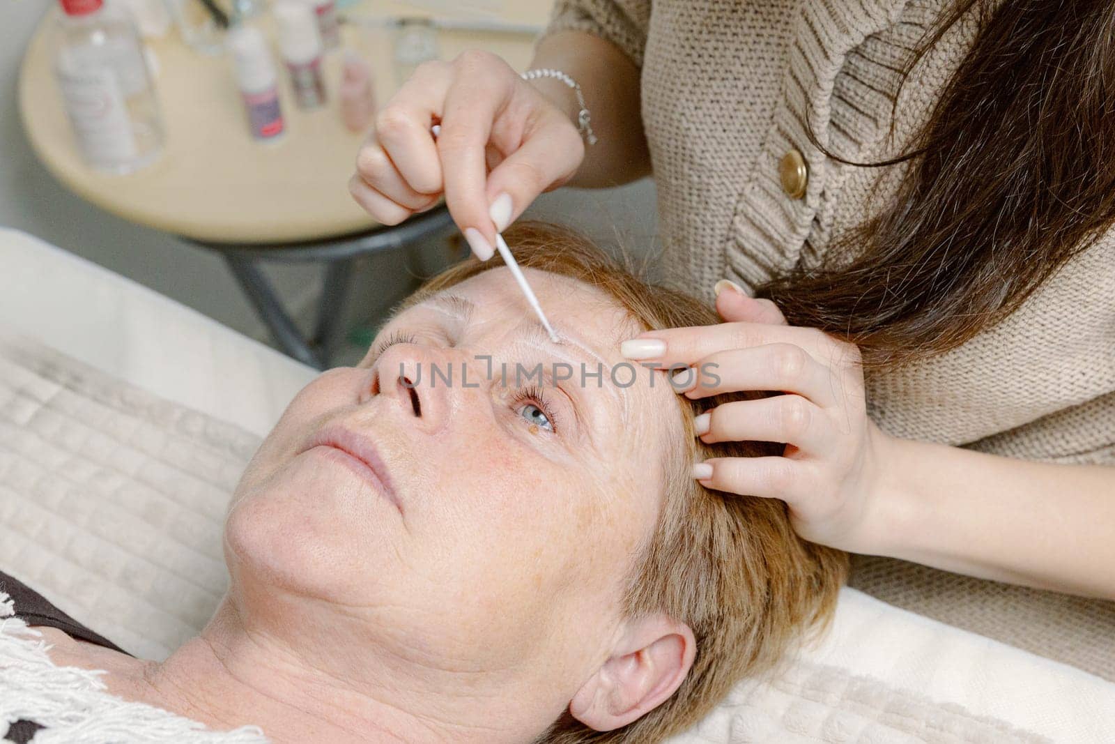 One young Caucasian young girl cosmetologist degreases the eyebrows of an elderly woman with closed eyes using a stick lying on a cosmetology bed in a home beauty salon, top side close-up view.