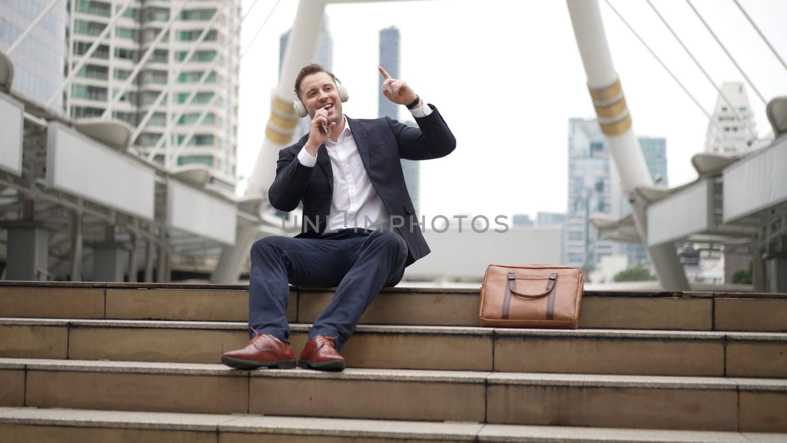 Professional business man listening headphone and using phone record voice while sitting at stair in urban city. Manager using headset listening relaxed song and moving along in lively mood. Urbane.