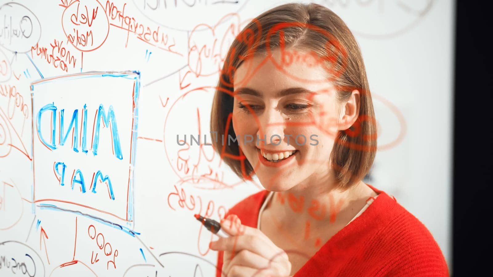 Closeup of young beautiful caucasian business leader presents marketing idea while writing marketing idea on glass board at creative meeting, brain storming discussing, working together. Immaculate.