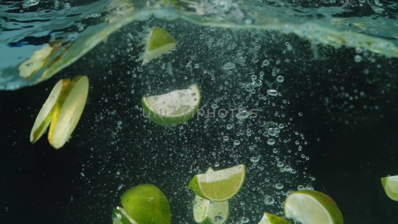 Macro of slices of lime being dropped into a glass of clear liquid. Bubbles rising up from the bottom of the glass. Close up of lime falling in to water. Garnished cocktail or decorated food. Pabulum.