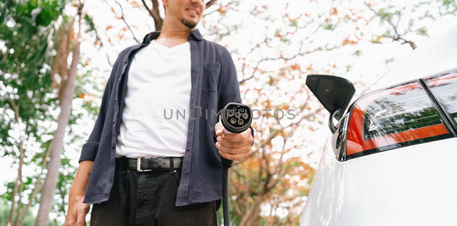 Panorama banner man recharging battery for electric car during autumnal road trip travel EV car in autumnal forest. Eco friendly travel on vacation during autumn with electric vehicle. Exalt