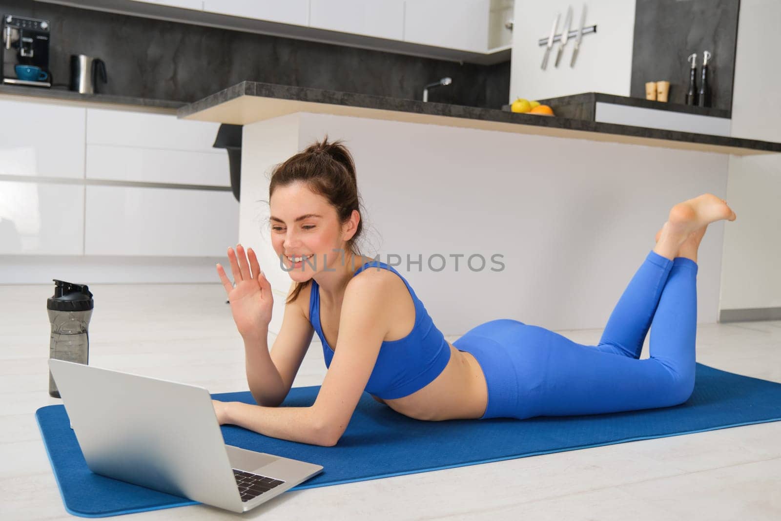 Portrait of happy young woman, doing fitness at home, connects to video chat on laptop, waves hand at computer to say hi, laying on yoga mat in activewear.