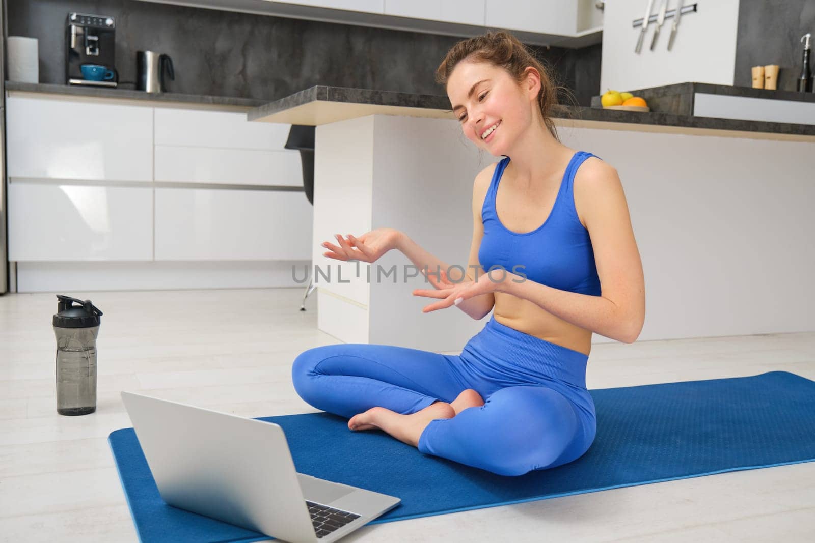 Image of young smiling woman, fitness instructor sitting on yoga mat, talking to client via online video chat on laptop, teaching aerobics, workout training.