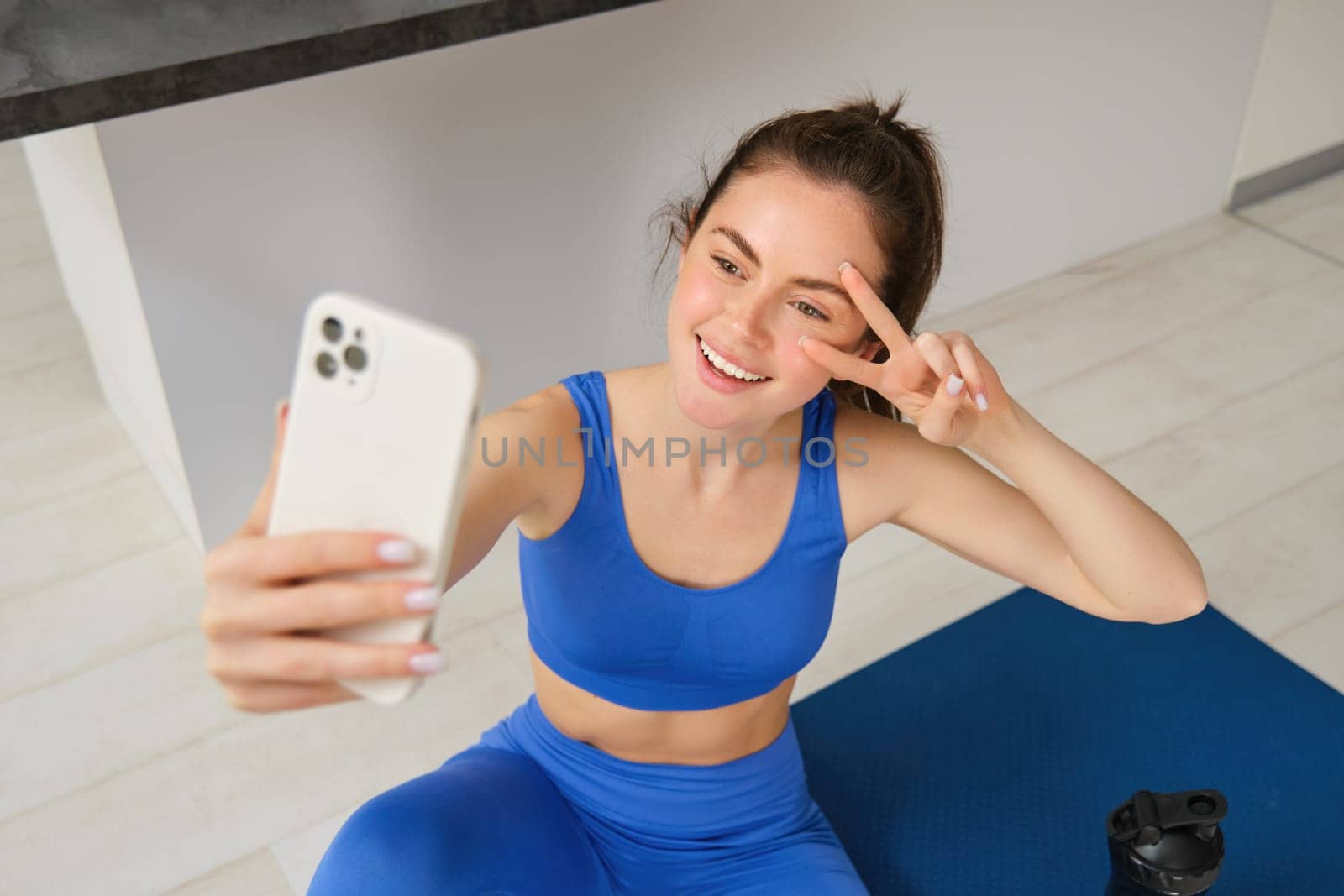 Portrait of beautiful female athlete, woman doing workout at home on rubber yoga mat, takes selfie on smartphone, makes photos for social media of fitness instructor.