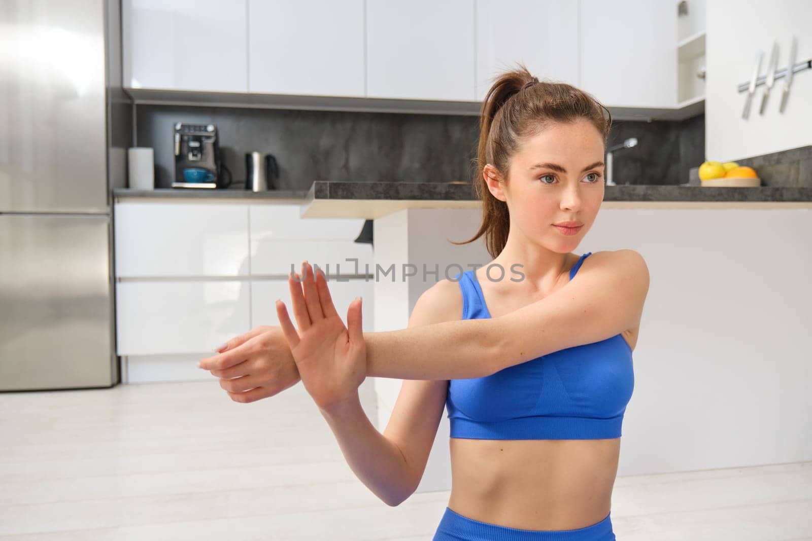 Image of fitness girl concentrates on workout, stretches hands before training session at home, follows online gym instructions.