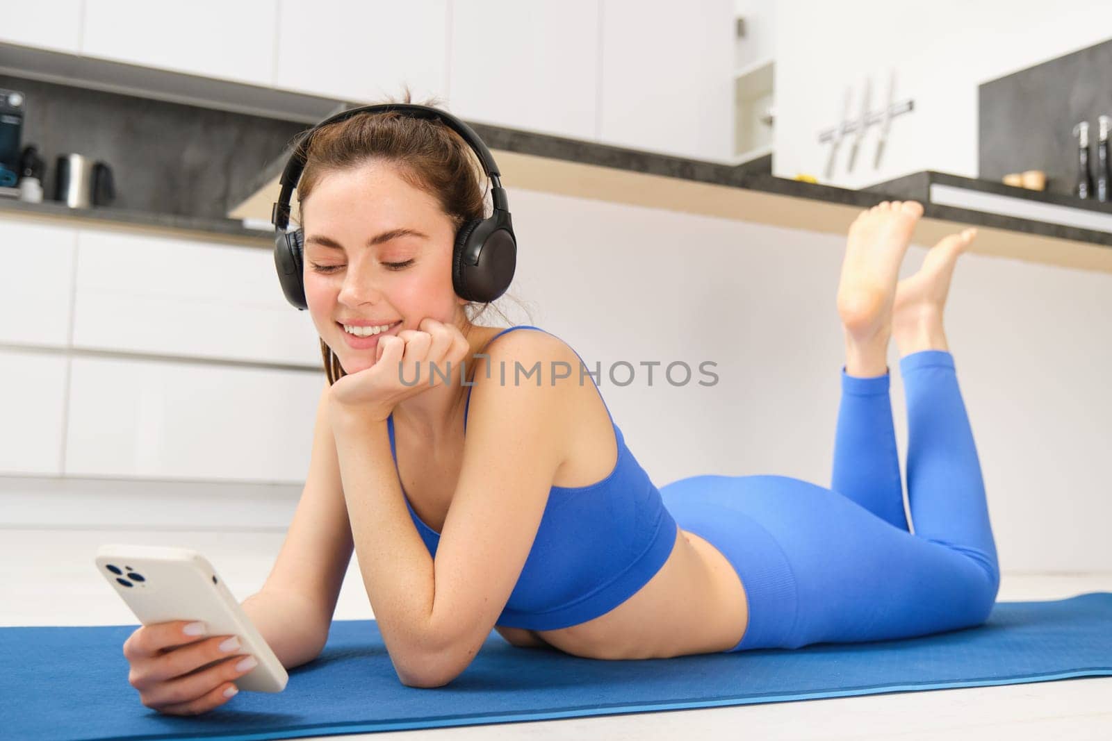 Portrait of fitness woman, listening yoga music in headphones, using her smartphone during workout at home, laying on blue sports mat.