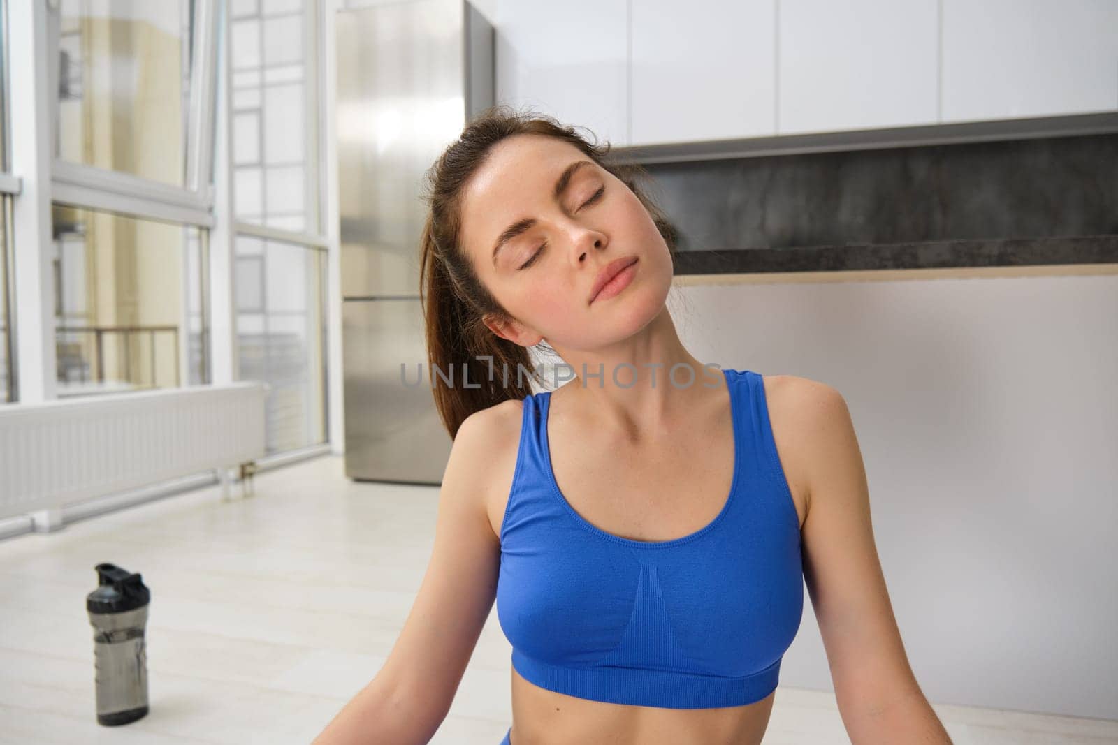 Portrait of young sportswoman, stretching her neck, warm-up before yoga exercises, doing fitness workout on rubber mat, wearing blue sportsbra and leggings.