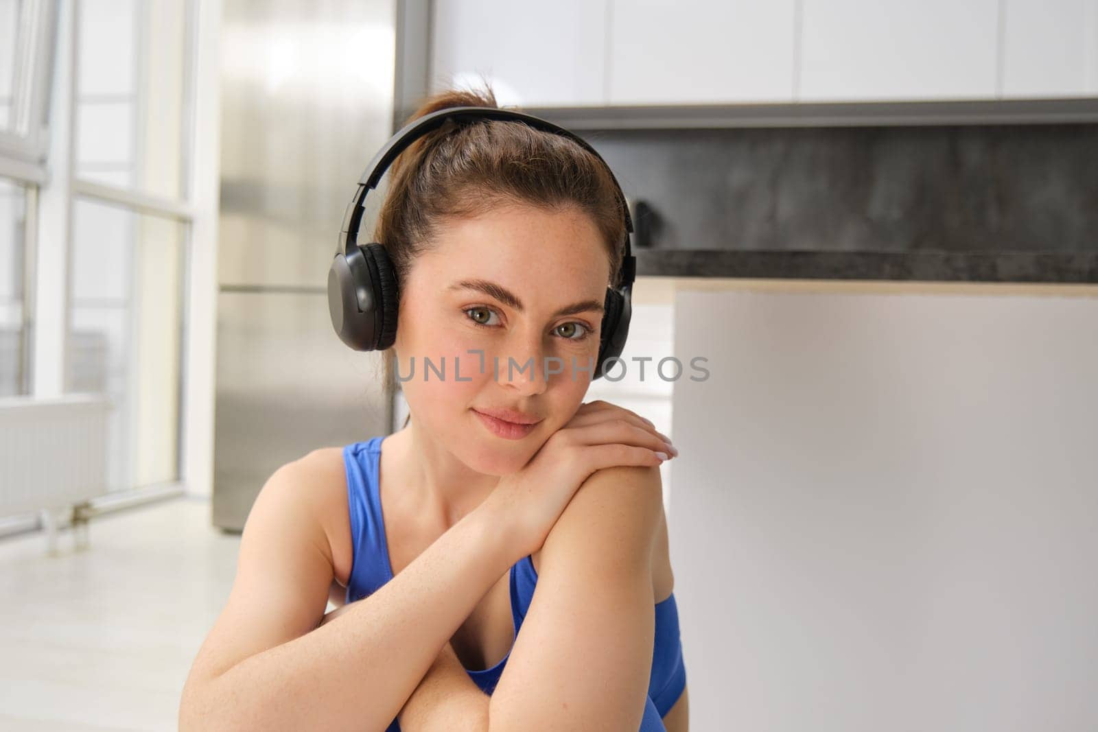 Close up portrait of beautiful brunette girl in sportswear, sits in wireless headphones and listens music, while doing workout training at home.