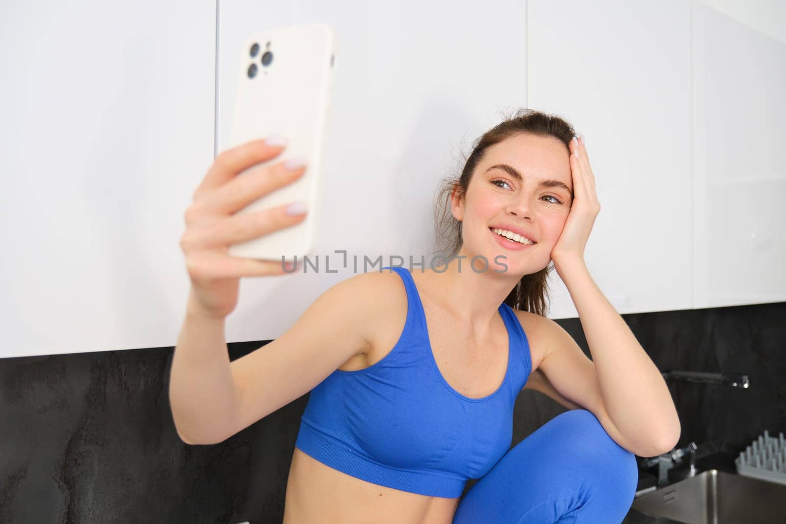 Image of fit and healthy, smiling fitness girl, posing for selfie on mobile phone, holding smartphone, looking at screen, taking photos in sportsbra and leggings.