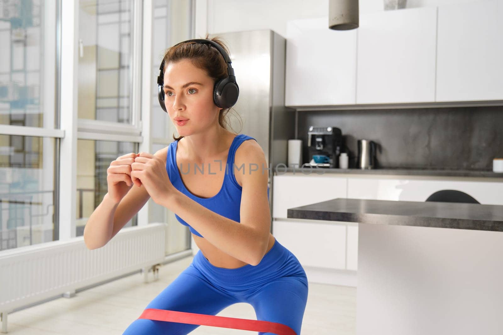 Portrait of woman doing squats with elastic resistance band, looking focused during workout, doing exercises at home.