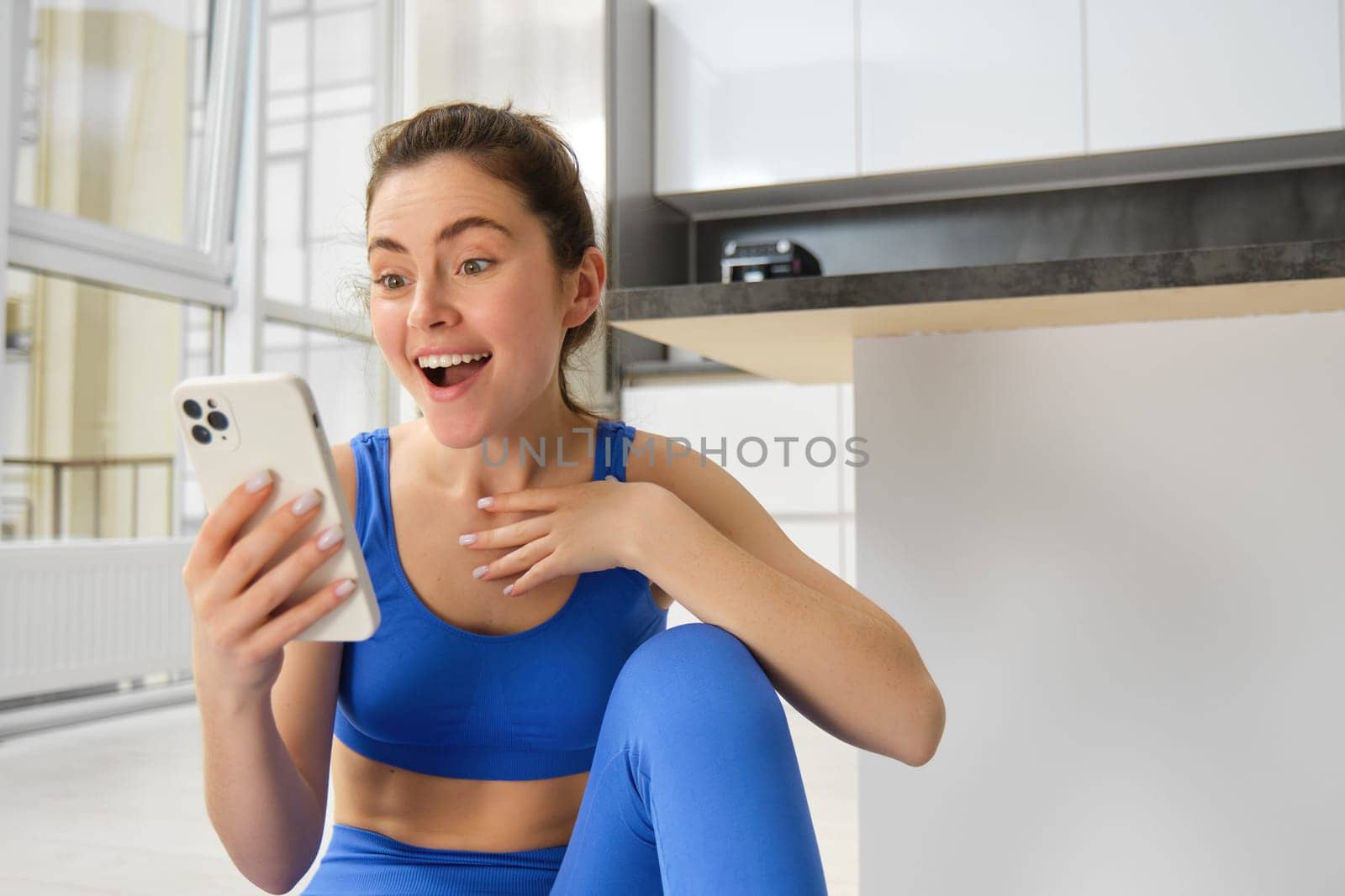 Indoor shot of surprised, happy young sportswoman looking at smartphone with smile and excitement, reading great news on phone message, sitting in blue sportsbra and leggings.