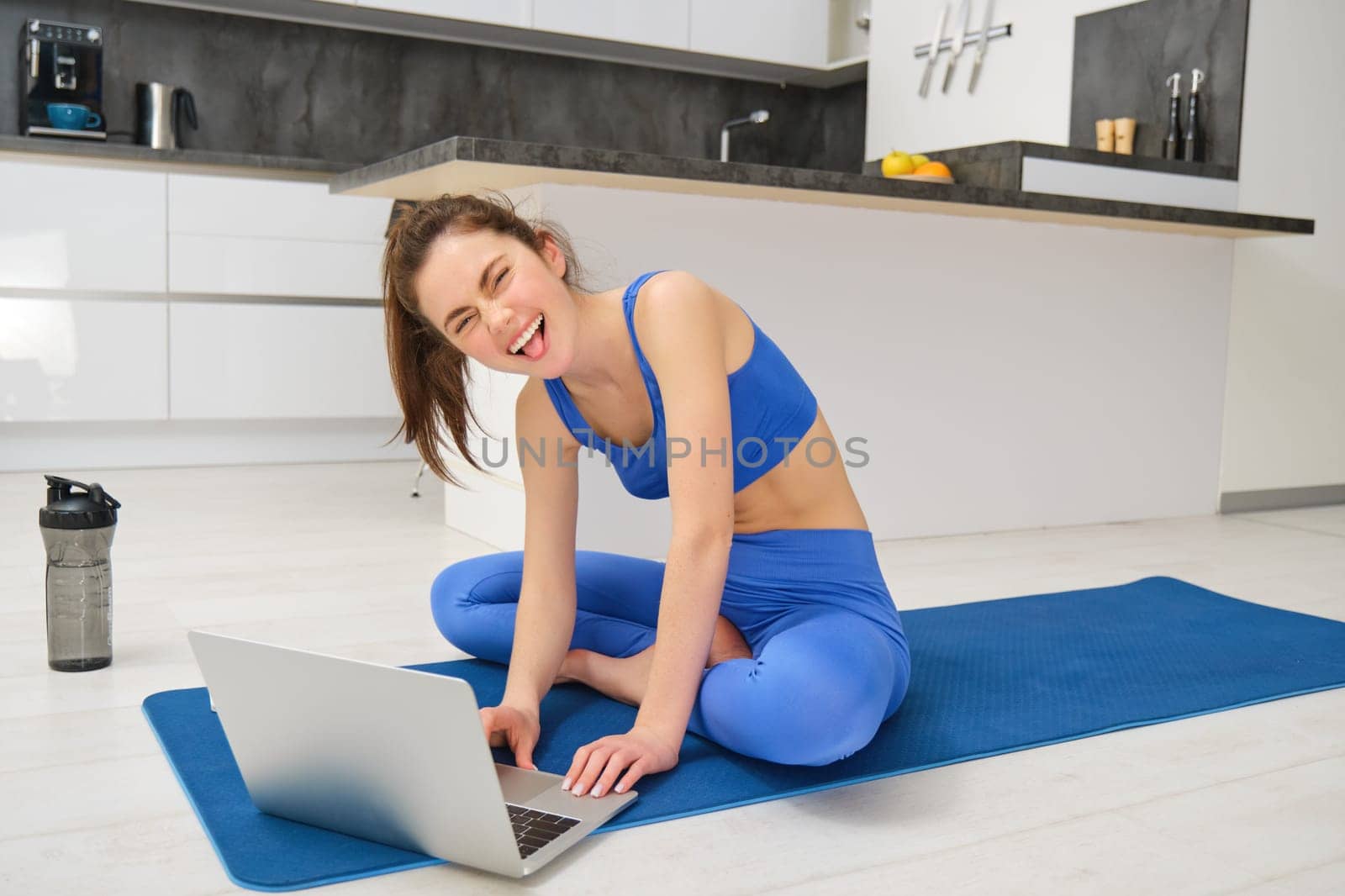 Portrait of happy young fitness woman, talking with client over laptop video chat, records her workout training session, showing aerobics exercises.