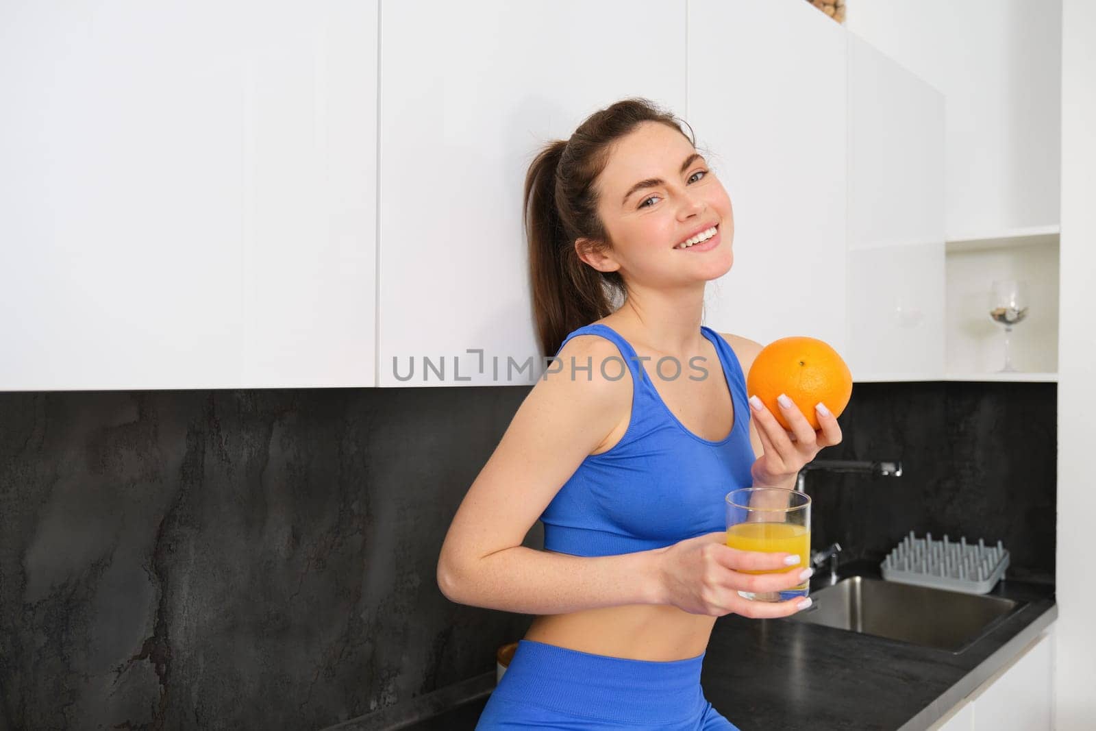 Indoor shot of woman after workout, standing in kitchen with fresh juice and an orange, drinking it.