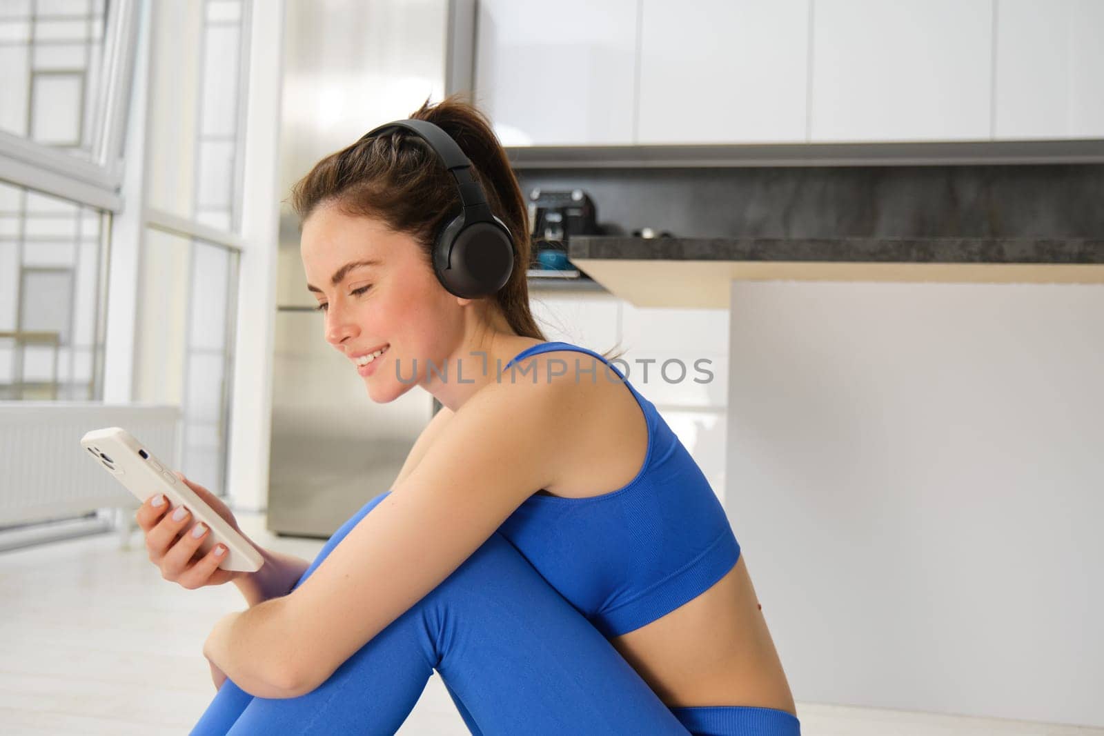 Portrait of brunette girl in fitness clothes, listens music in wireless headphones, holds smartphone, workout from home, looking at training video instructions.