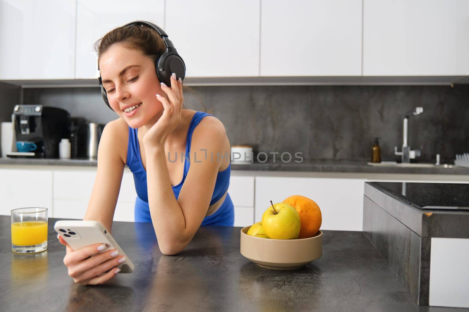 Image of happy, stylish young sports woman, standing in kitchen and drinking orange juice, listening music in headphones, using smartphone app.