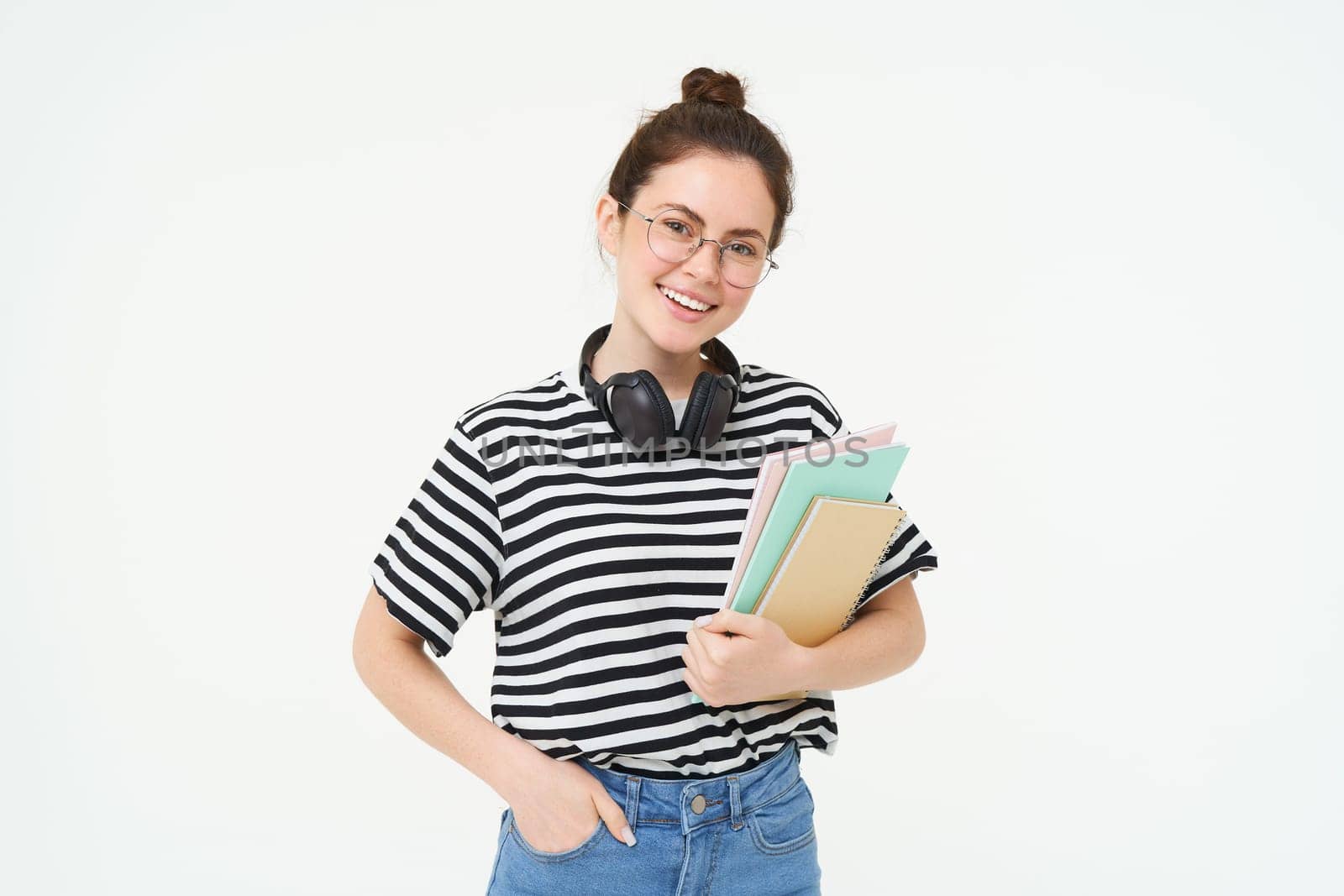 Young woman with notebooks, books and study material, posing over white background, wears headphones over neck.