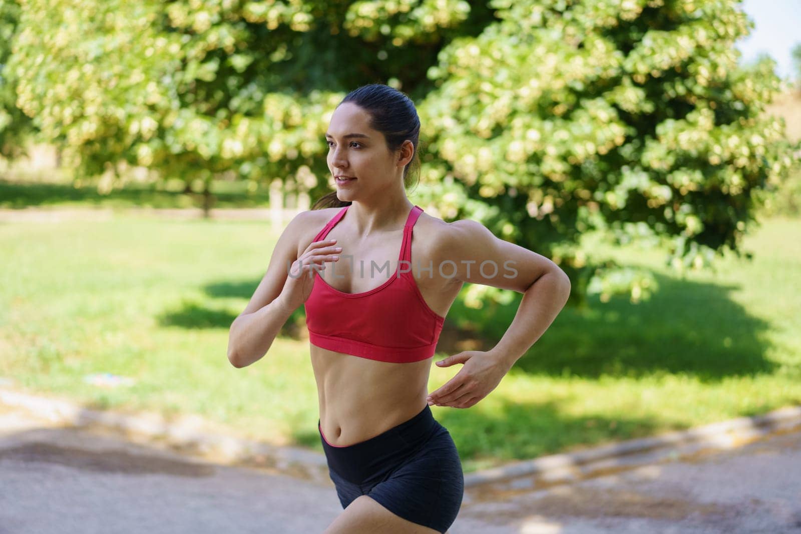 A young woman in red sports bra and black shorts is jogging in a green park on a sunny day, promoting fitness and a healthy lifestyle, embodying keywords like active, exercise, health, and wellness