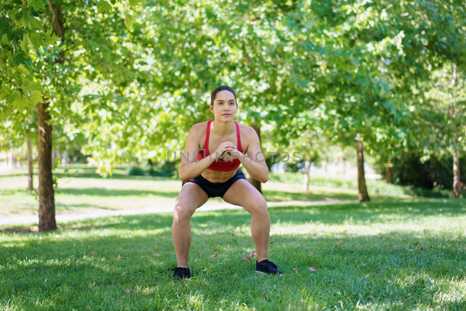 A woman in sportswear is squatting in a sunny park, promoting outdoor fitness, strength training, and a healthy lifestyle for her wellbeing. She highlights active living and dedication to exercise