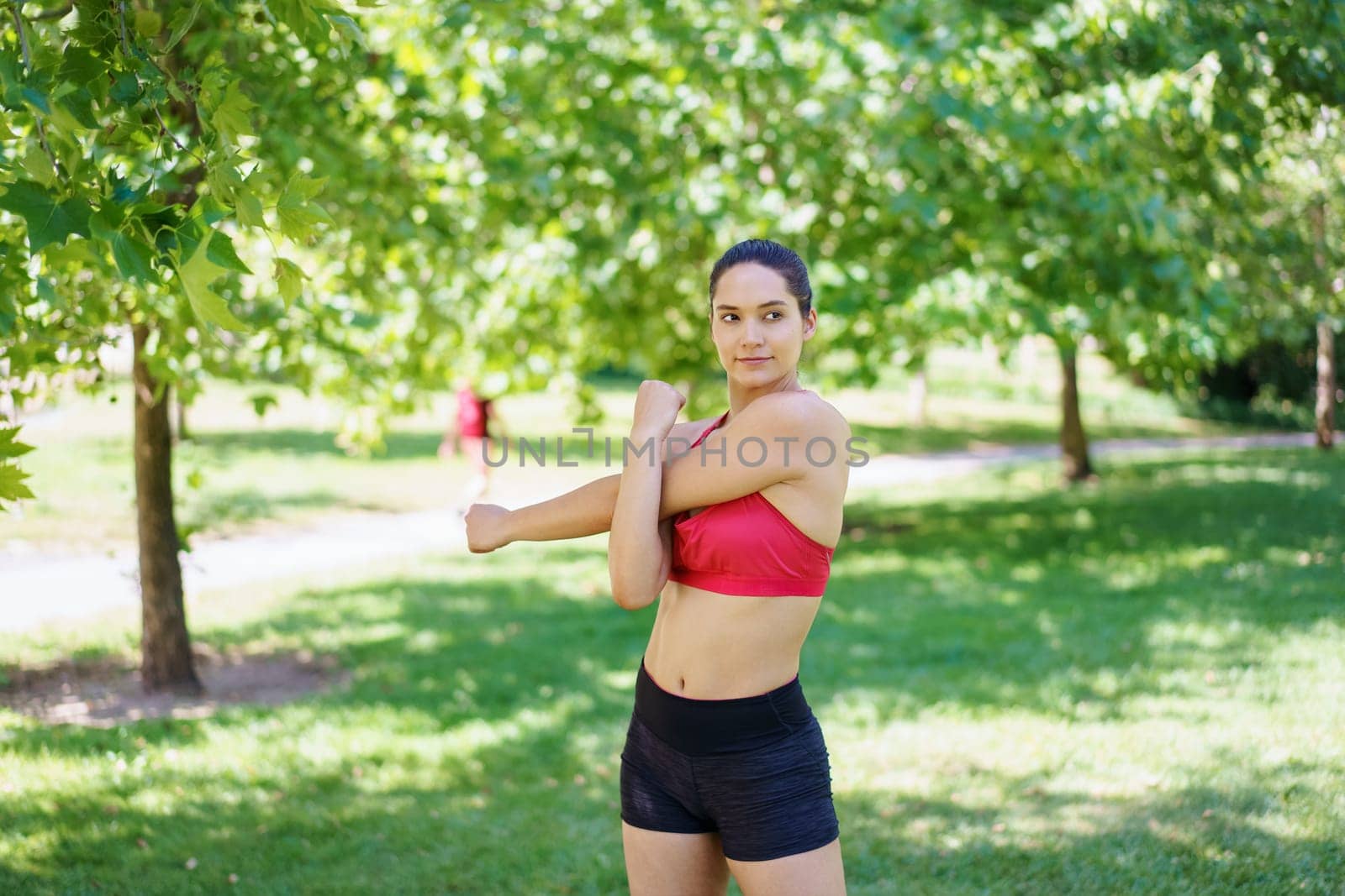 In a lush green park, a young athletic woman in red sports bra and black shorts is stretching her arm, getting ready for outdoor workout, exemplifying dedication to fitness and strength