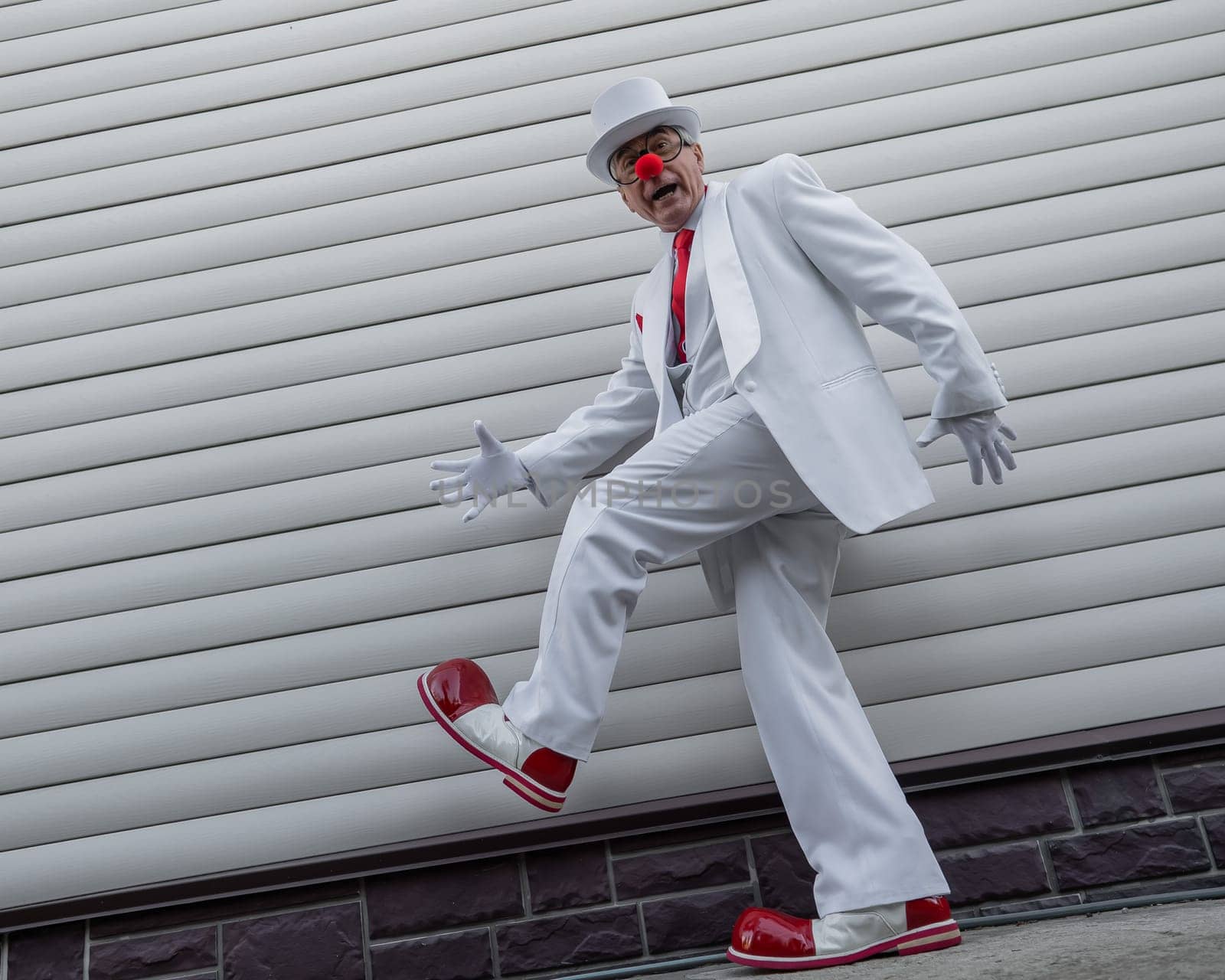 An elderly man in a white suit, huge boots and a clown nose walks funny. by mrwed54