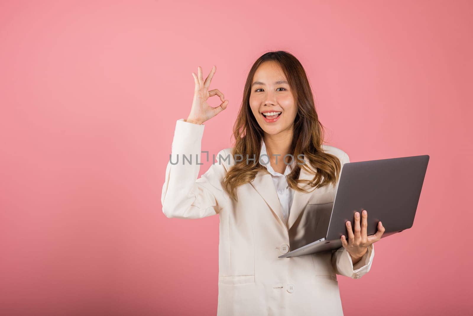 Portrait business woman smiling she holding laptop computer and show OK gesture isolated pink background, Asian female showing ok-sign by Sorapop